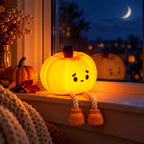 A glowing orange jack-o'-lantern window light with a sad face and rope legs sits on a windowsill at night, surrounded by seasonal decor including a small pumpkin, dried flowers, and a knitted blanket.