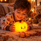 A young boy wearing patterned pajamas lies on a carpeted floor, smiling at a glowing orange pumpkin-shaped night light with legs and a sad face. The room has warm string lights and a teddy bear in the background.