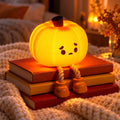A glowing orange pumpkin-shaped lamp with a sad face and rope legs, resting on a textured surface in a warm, dimly lit setting.